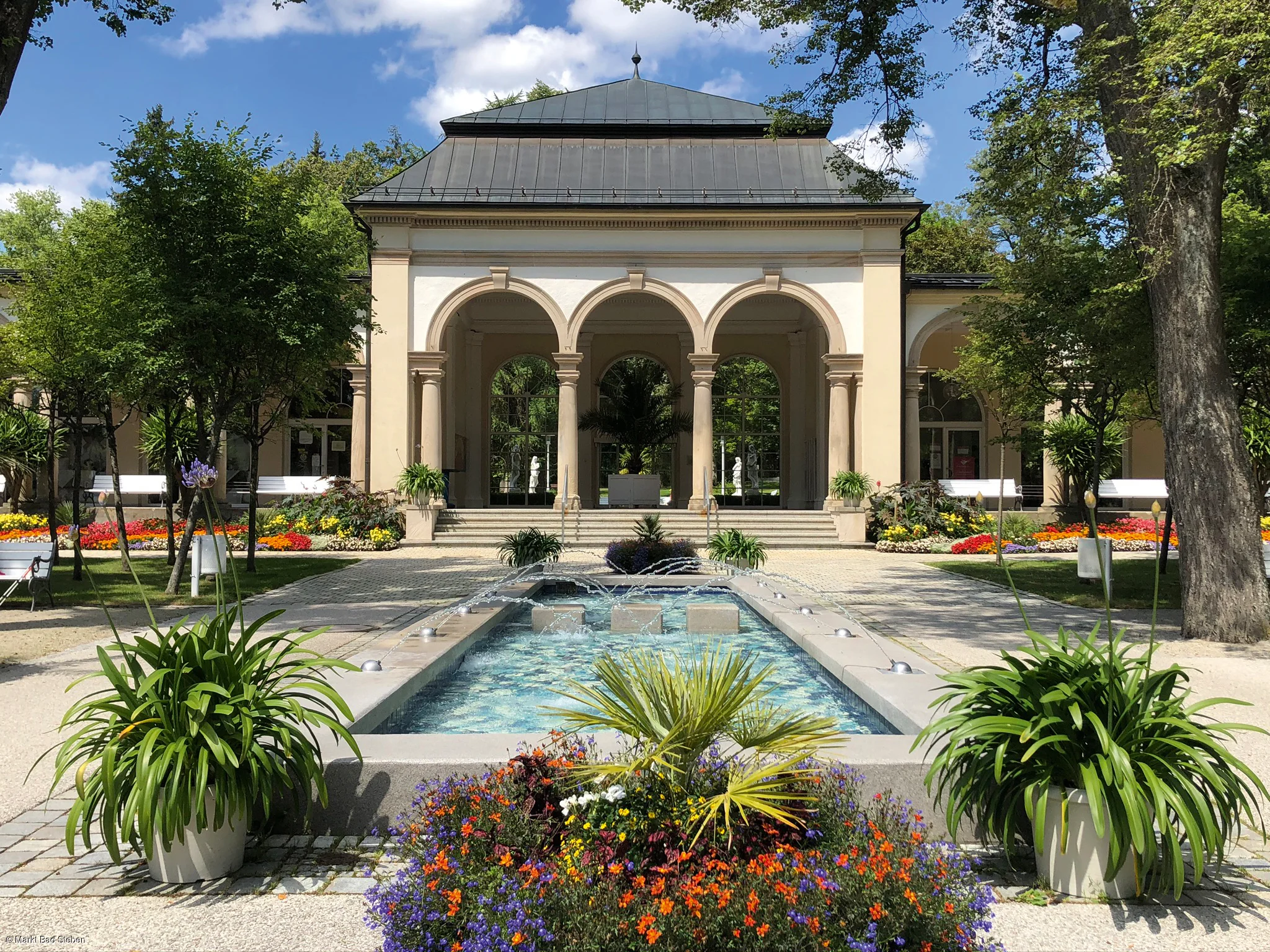 Gebäude mit Arkaden, davor ein Springbrunnen und Blumenbeete in einem Park bei sonnigem Wetter.