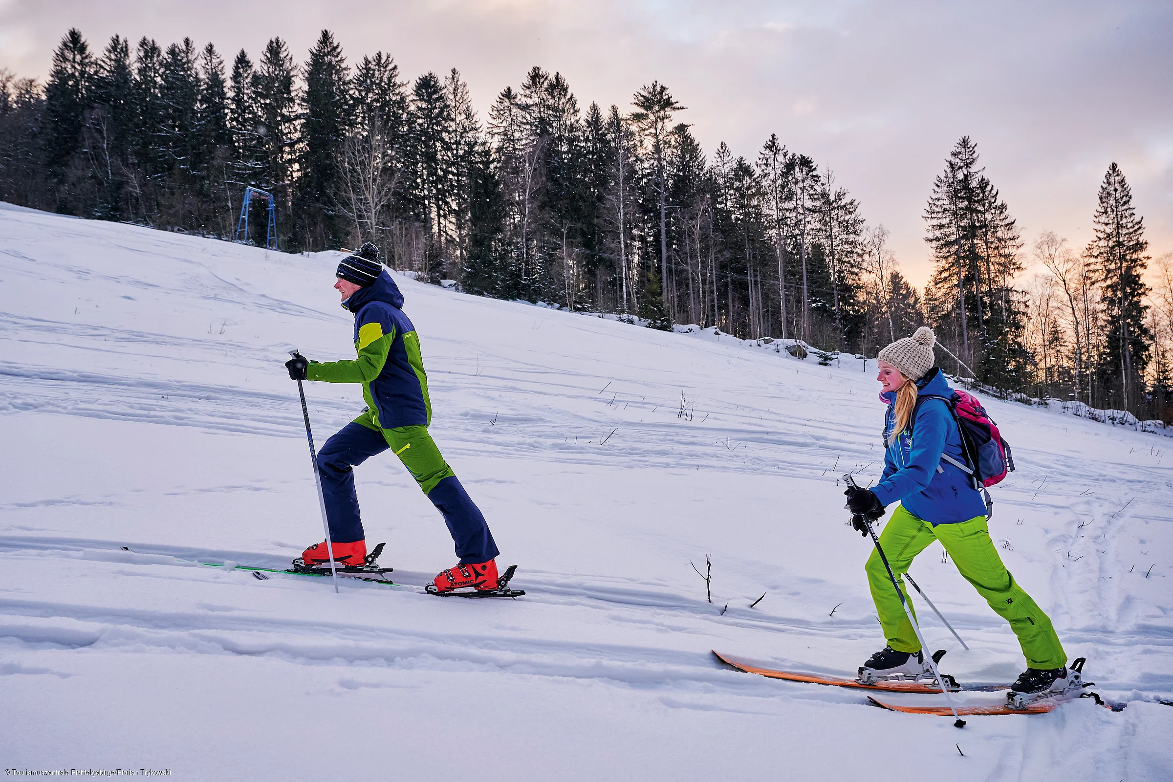 Zwei Personen auf schneebedecktem Hang vor Wald mit Abendhimmel.
