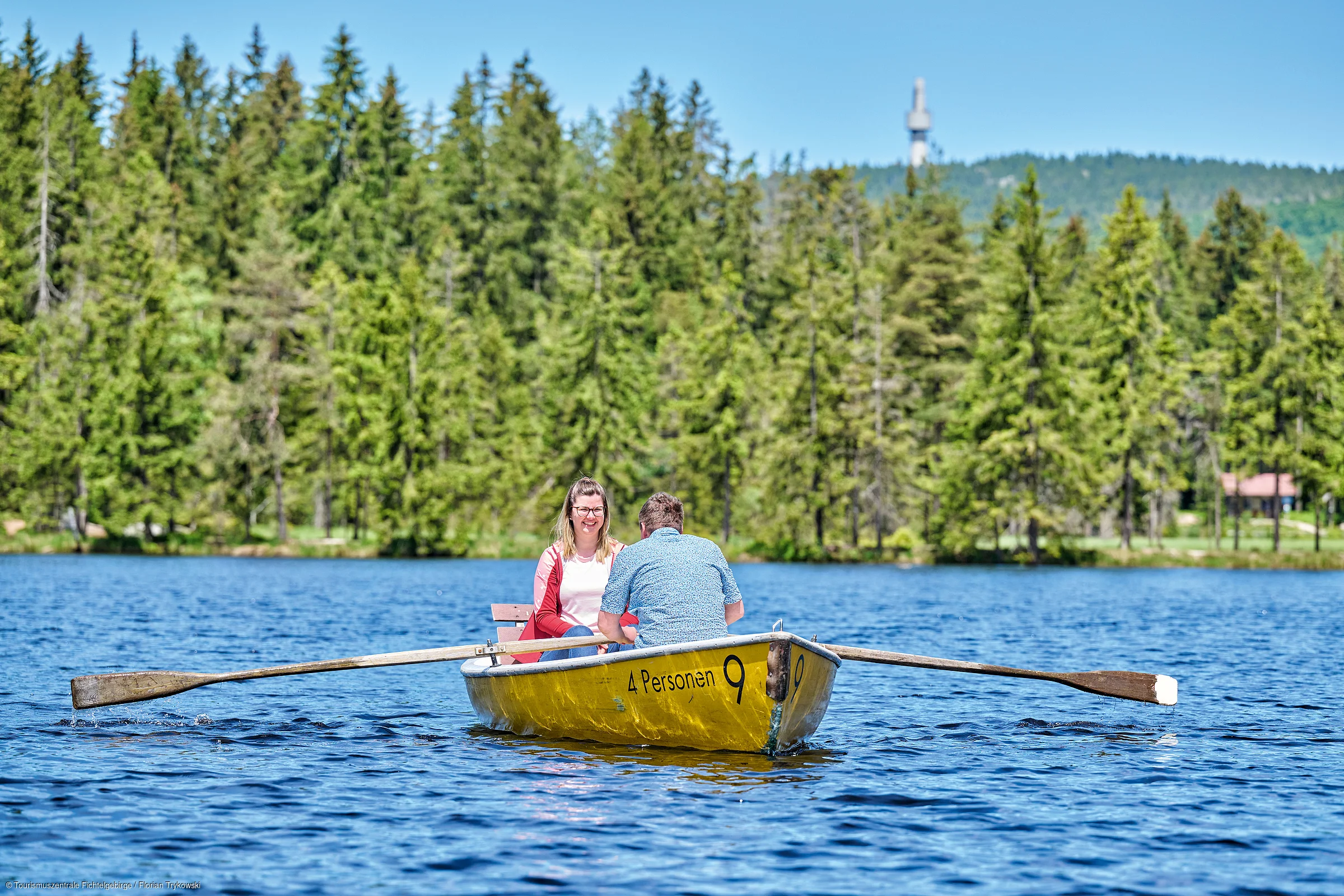 Zwei Personen sitzen in einem gelben Ruderboot auf einem See vor einem bewaldeten Ufer.