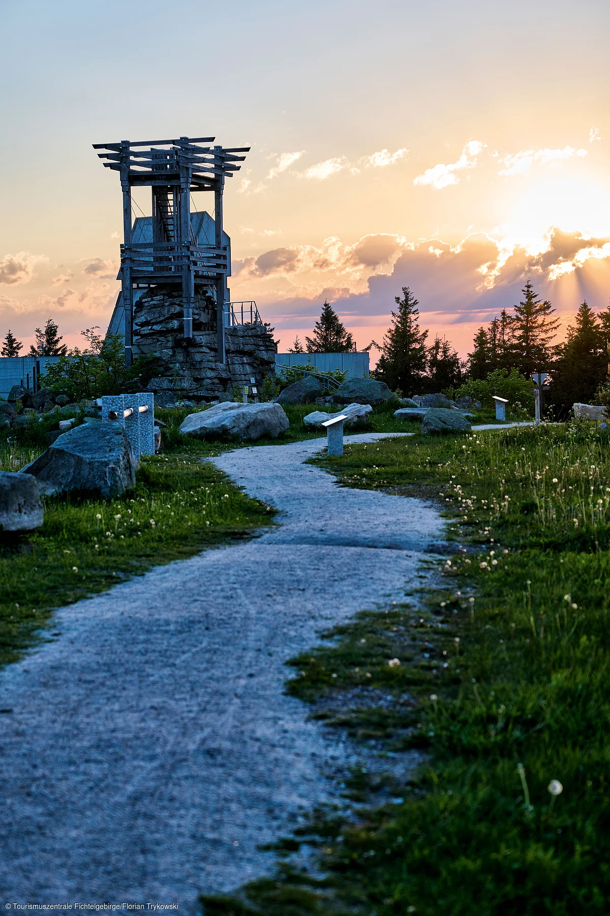 Wanderweg mit Steinen und Holz-Aussichtsturm bei Sonnenuntergang, umgeben von Wiesen und Bäumen.