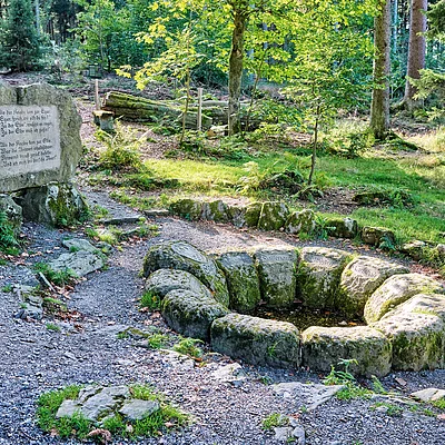 Kreisförmige Steinanordnung mit Wasser in einem Wald, daneben großer Stein mit Inschrift und Waldweg.