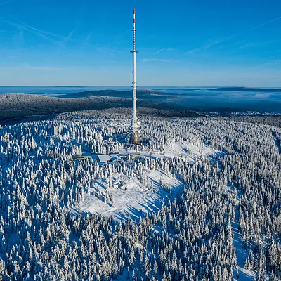 Luftaufnahme eines mit Schnee bedeckten Waldes mit einem hohen Sendemast in der Mitte bei klarem Himmel.
