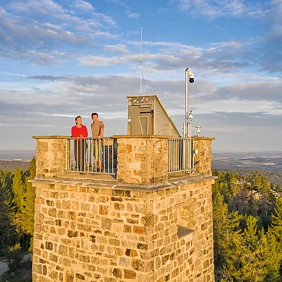Zwei Personen stehen auf einer Aussichtsplattform eines steinernen Turms mit Wald und Himmel im Hintergrund.