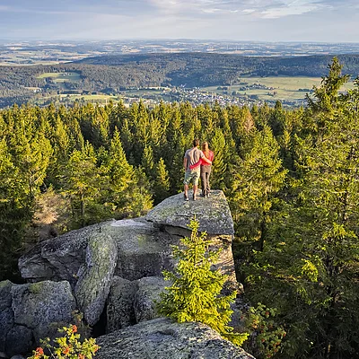 Zwei Personen stehen auf einem Felsen und blicken auf einen Wald und eine Landschaft mit Dörfern und Feldern.