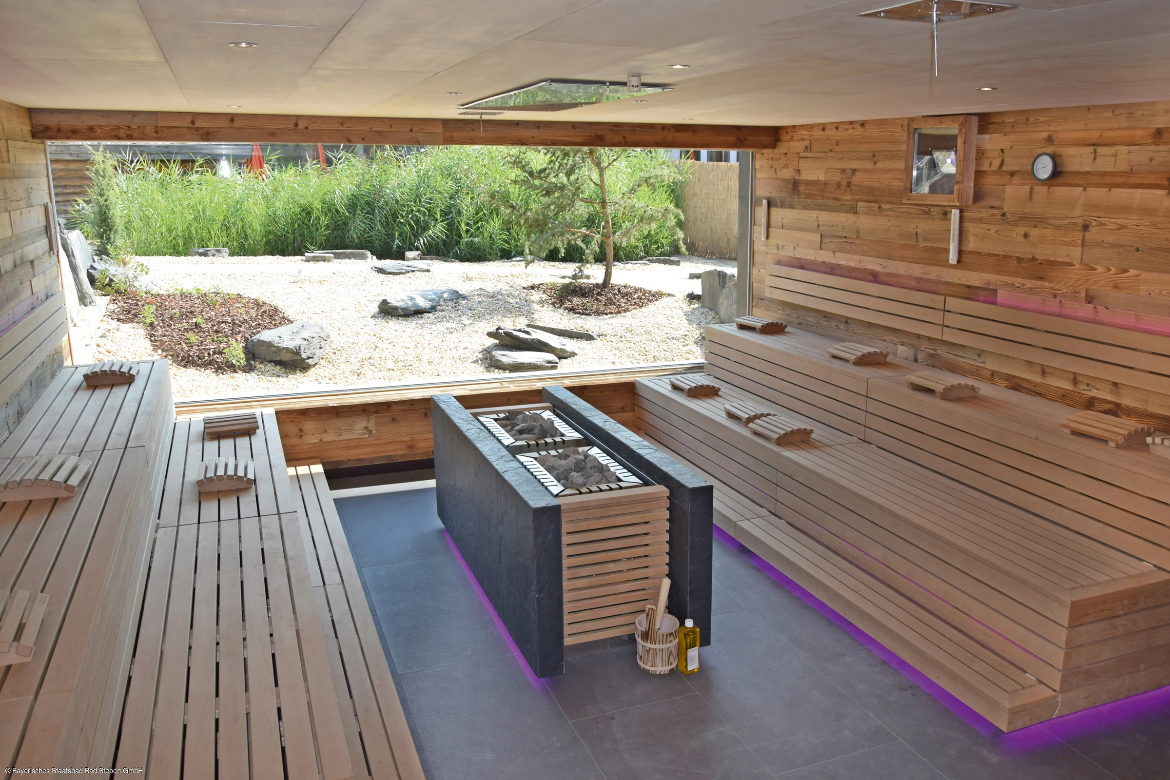 Innenraum einer Sauna mit Holzbänken, zentralem Ofen mit Steinen und großem Fenster zum Gartenbereich
