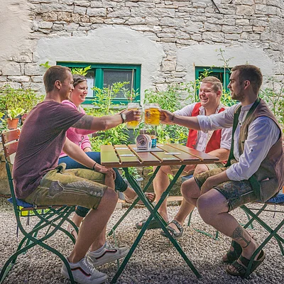 Vier Personen sitzen an einem Tisch im Freien und stoßen mit Bierkrügen an. Im Hintergrund eine Steinmauer.