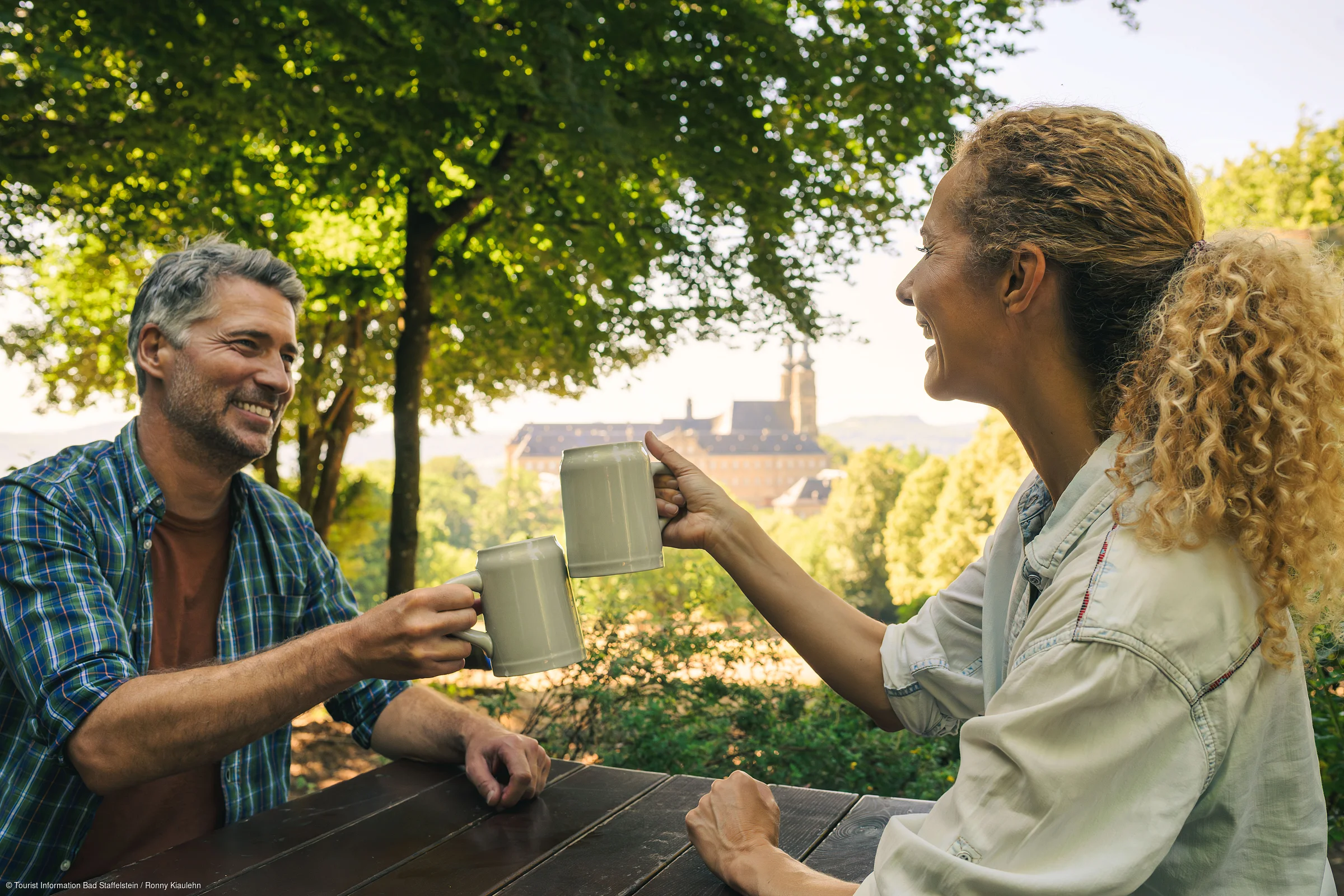 Zwei Personen stoßen mit Bierkrügen an einem Holztisch im Freien an, im Hintergrund Bäume und ein Gebäude.