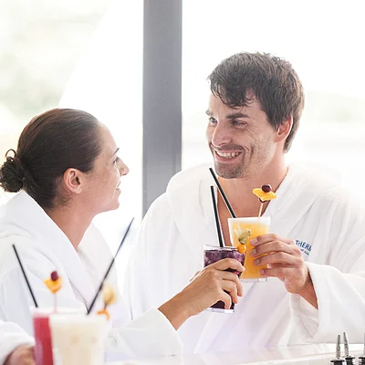 Zwei Personen in weißen Bademänteln stoßen mit bunten Getränken an einer Bar an.