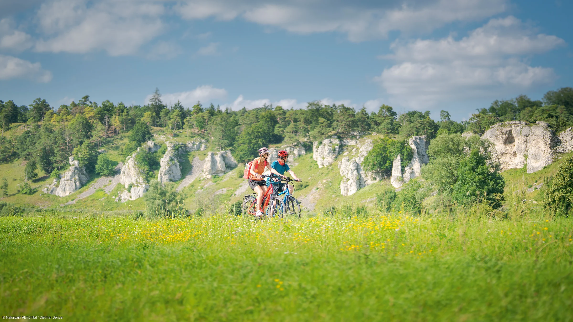 Zwei Personen fahren mit Fahrrädern durch eine grüne Wiese, im Hintergrund Felsen und Bäume.