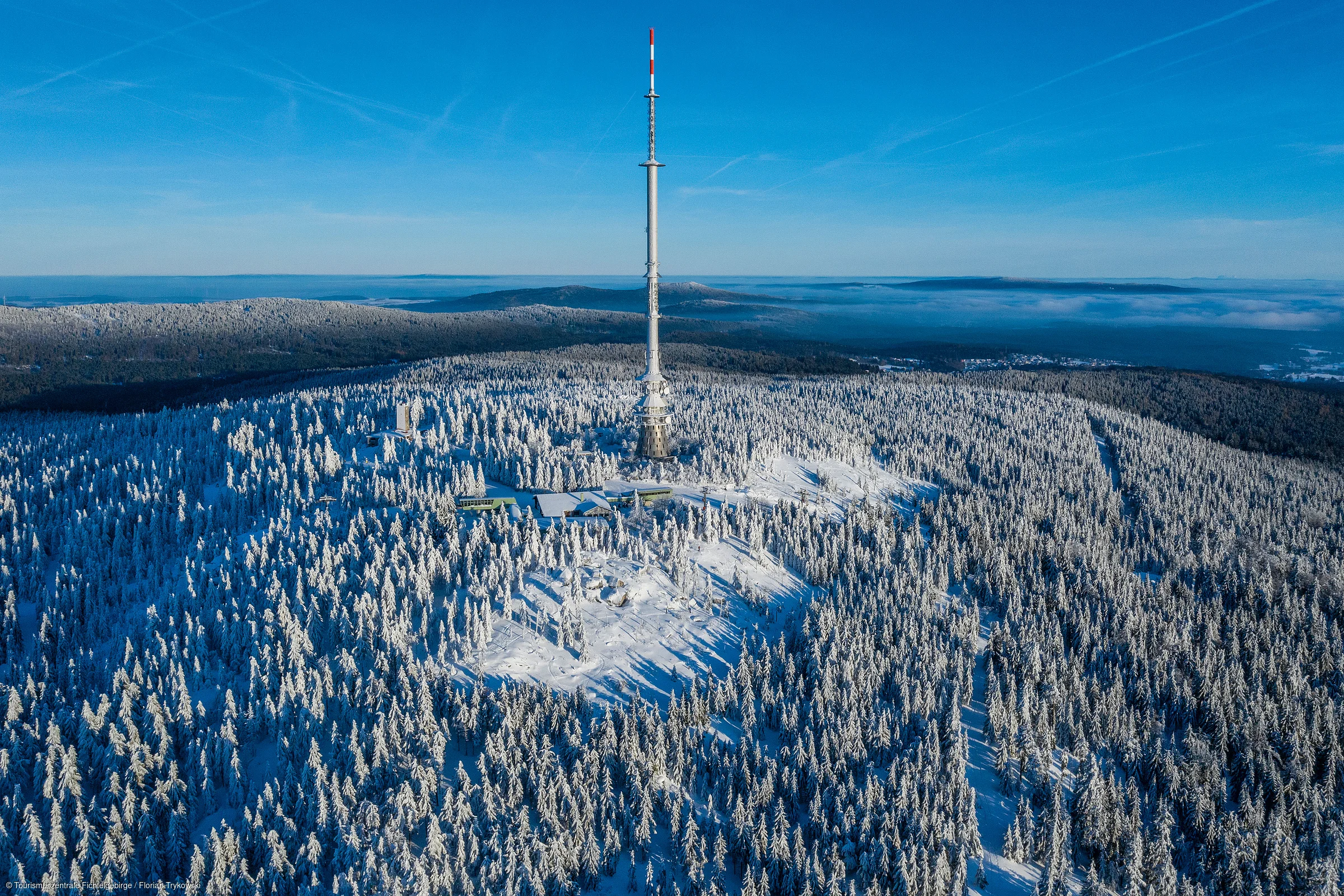 Luftaufnahme eines mit Schnee bedeckten Waldes mit einem hohen Sendemast in der Mitte bei klarem Himmel.