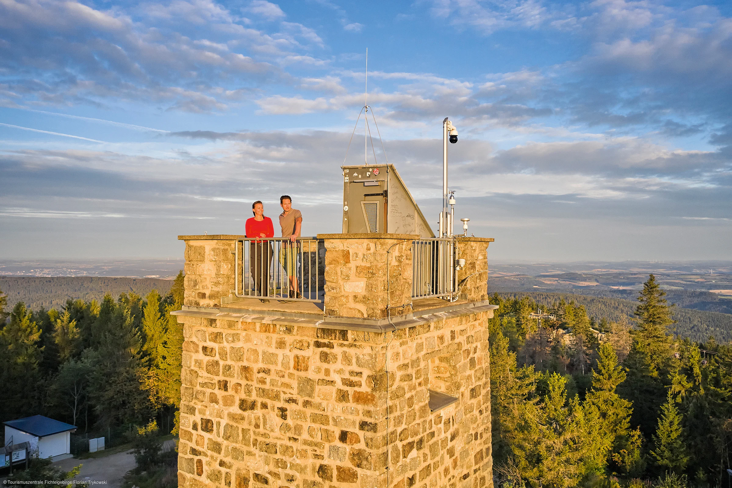 Zwei Personen stehen auf einer Aussichtsplattform eines steinernen Turms mit Wald und Himmel im Hintergrund.