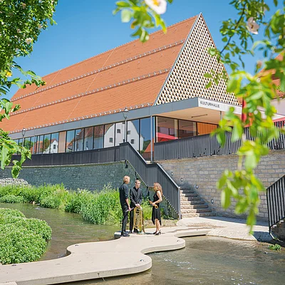 Eine moderne Halle mit der Aufschrift „Kulturhalle“ neben einem Fluss. Drei Personen mit Musikinstrumenten stehen am Ufer.