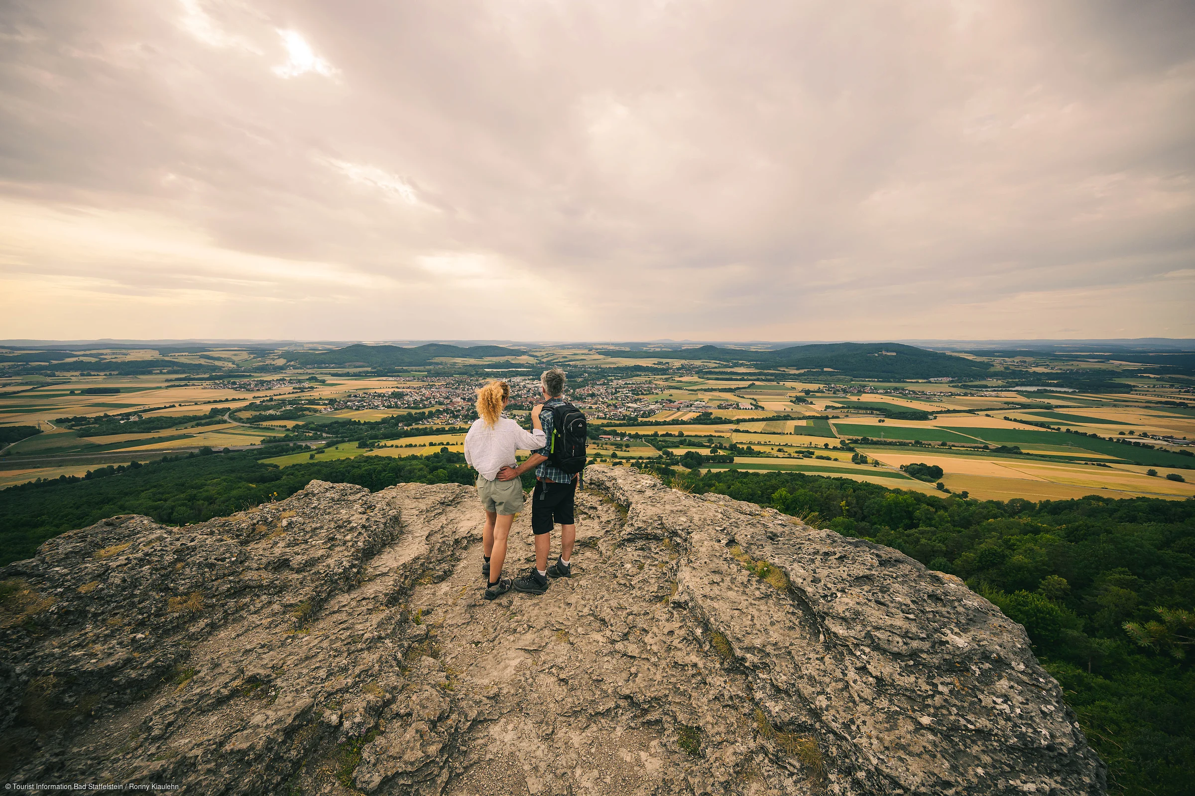 Zwei Wanderer stehen auf einem Felsen und blicken auf eine weite Landschaft mit Feldern und Wäldern.