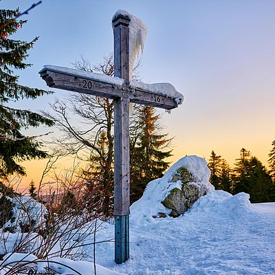 Holzkreuz mit Schnee bedeckt im verschneiten Wald bei Sonnenuntergang, im Hintergrund Tannenbäume und Felsen.