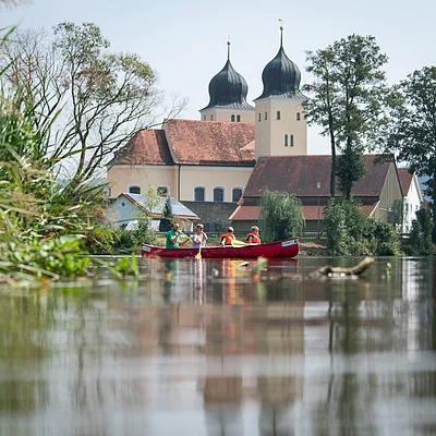 Zwei Personen in einem roten Kanu auf einem Fluss, im Hintergrund eine Kirche mit Zwiebeltürmen.