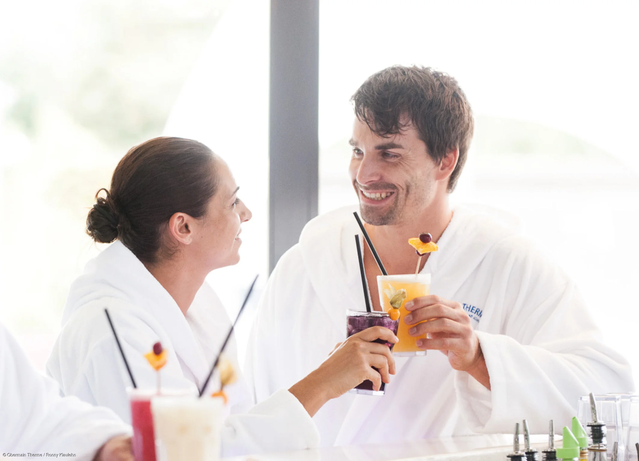 Zwei Personen in weißen Bademänteln stoßen mit bunten Getränken an einer Bar an.