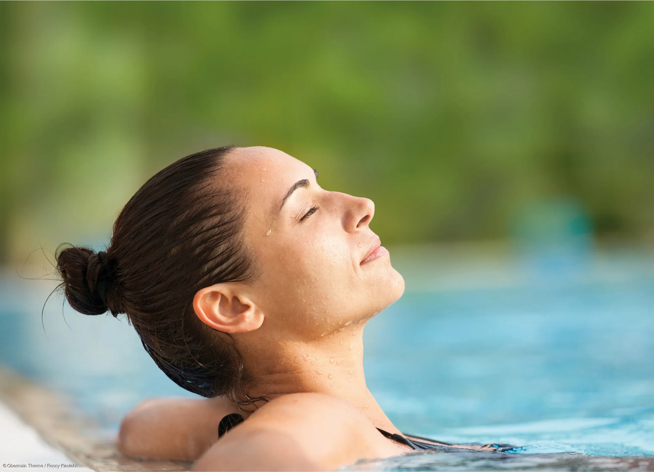 Frau mit nassem Haar im Dutt entspannt im Schwimmbecken mit geschlossenen Augen und Wasser im Hintergrund.