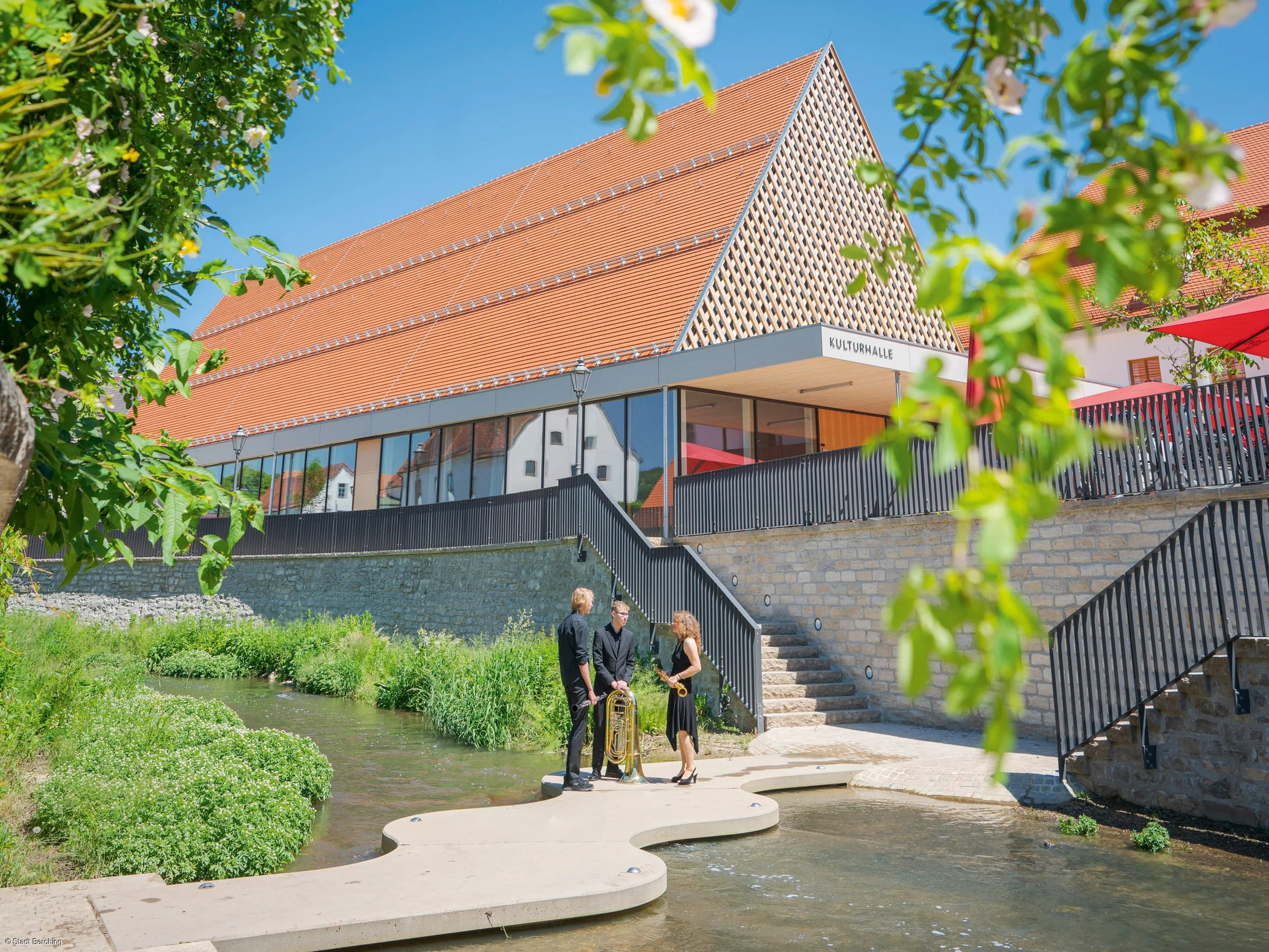 Eine moderne Halle mit der Aufschrift „Kulturhalle“ neben einem Fluss. Drei Personen mit Musikinstrumenten stehen am Ufer.