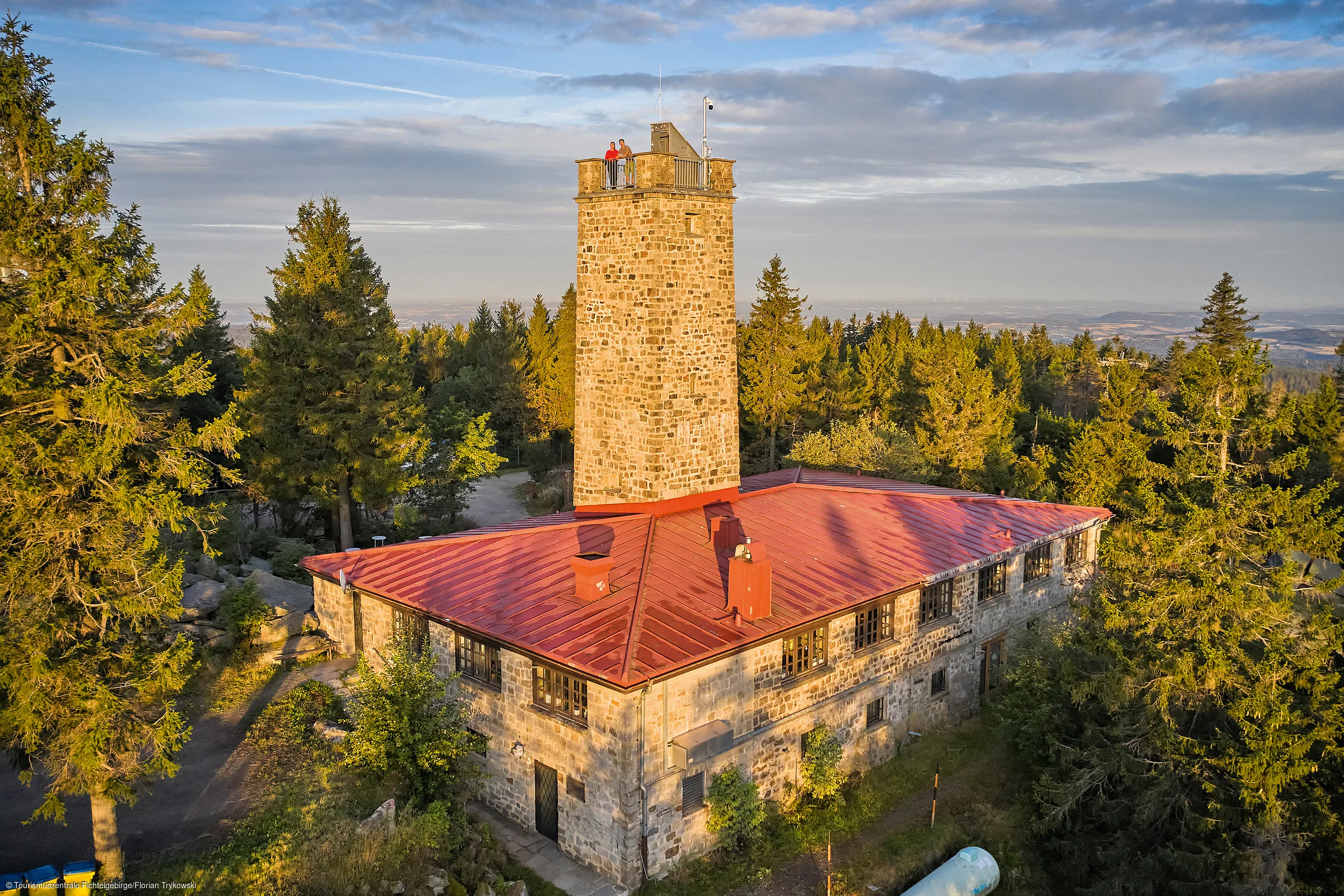 Steinerner Aussichtsturm mit rotem Dach umgeben von Nadelbäumen bei bewölktem Himmel.