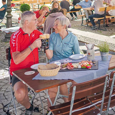 Ein älteres Paar sitzt an einem Tisch im Freien, stößt mit Bierkrügen an und hat eine Brotzeitplatte vor sich.