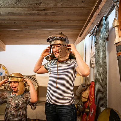 Kinder probieren antike Helme und Kettenhemden in einem Raum mit historischen Waffen an.