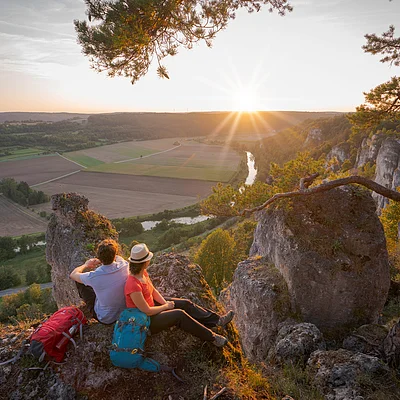 Zwei Wanderer sitzen auf Felsen mit Rucksäcken und blicken auf ein Tal mit Fluss und Sonnenuntergang.