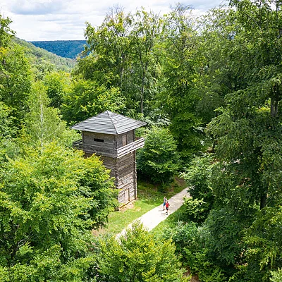 Holzturm neben Wanderweg im dichten grünen Wald mit zwei Personen auf dem Weg sichtbar