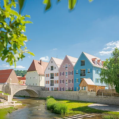 Bunte Häuserreihe an einem Fluss, mit einer Steinbrücke im Vordergrund und einem Kirchturm im Hintergrund.
