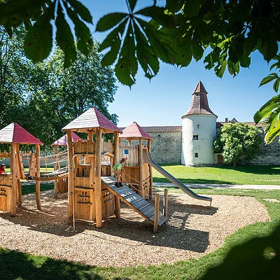 Spielplatz mit Holzklettergerüst, Rutsche und Seilbrücke, im Hintergrund ein historischer Turm.