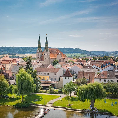 Stadtansicht mit Kirche, Wohnhäusern und Fluss im Vordergrund, umgeben von grüner Landschaft.