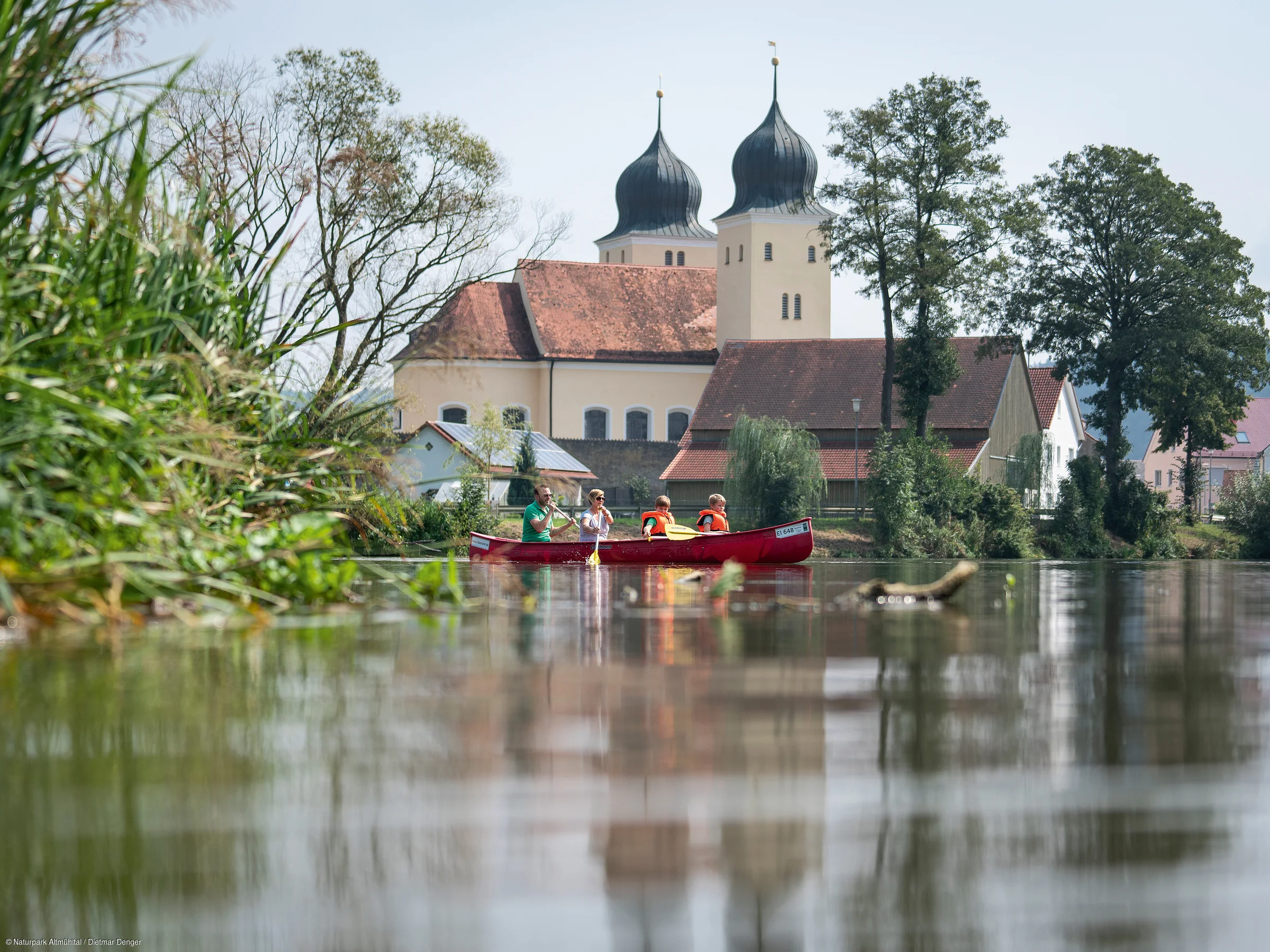 Zwei Personen in einem roten Kanu auf einem Fluss, im Hintergrund eine Kirche mit Zwiebeltürmen.