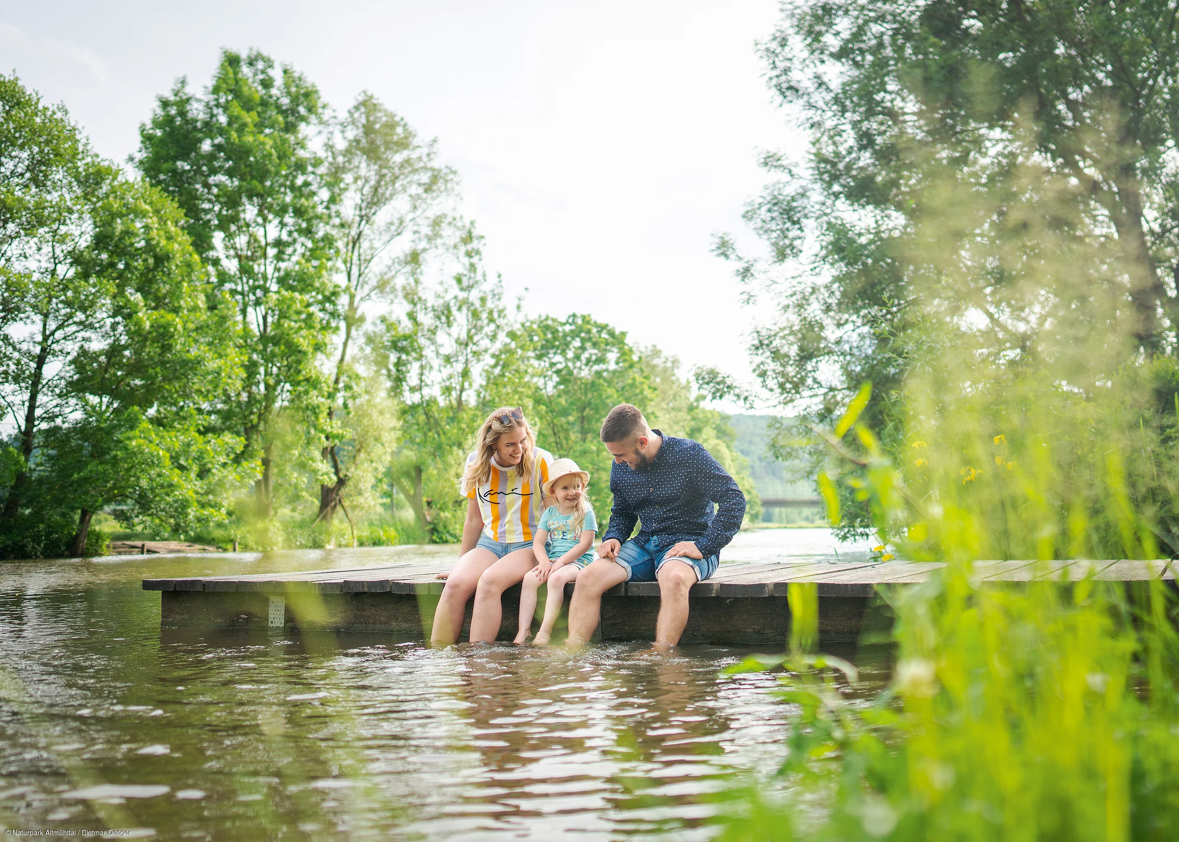 Eine Familie sitzt auf einem Steg am Wasser, umgeben von Bäumen und Pflanzen.