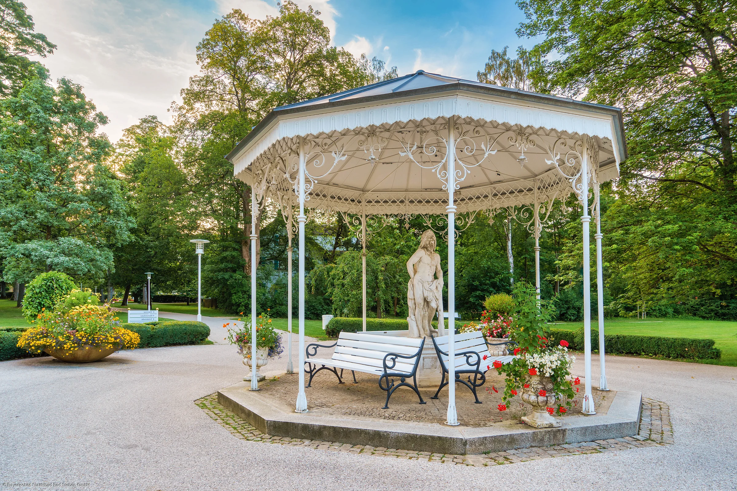 Weißes Pavillon mit Statue und Bänken in einem Park mit Bäumen und Blumenarrangements