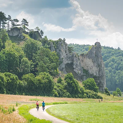 Zwei Radfahrer auf einem Weg durch eine grüne Landschaft mit Felsen und Bäumen im Hintergrund.