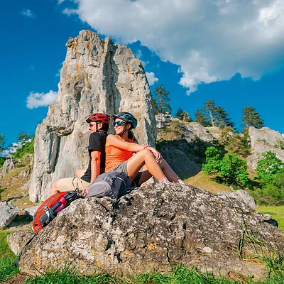 Zwei Personen mit Fahrradhelmen sitzen Rücken an Rücken auf einem Felsen vor einer Felsformation und blauem Himmel.