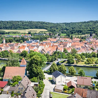 Stadtansicht mit Fluss, Brücke und roten Dächern, umgeben von Bäumen und Hügeln im Hintergrund.