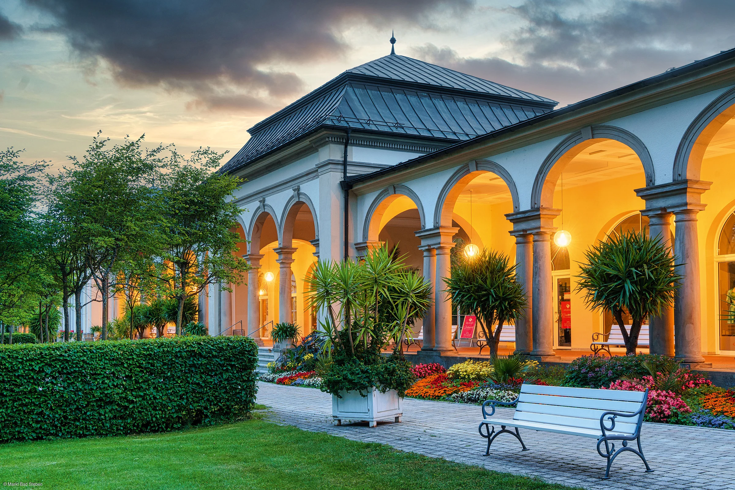 Gebäude mit Arkaden, beleuchteten Bögen, Blumenbeeten, Bänken und Bäumen bei Abenddämmerung.