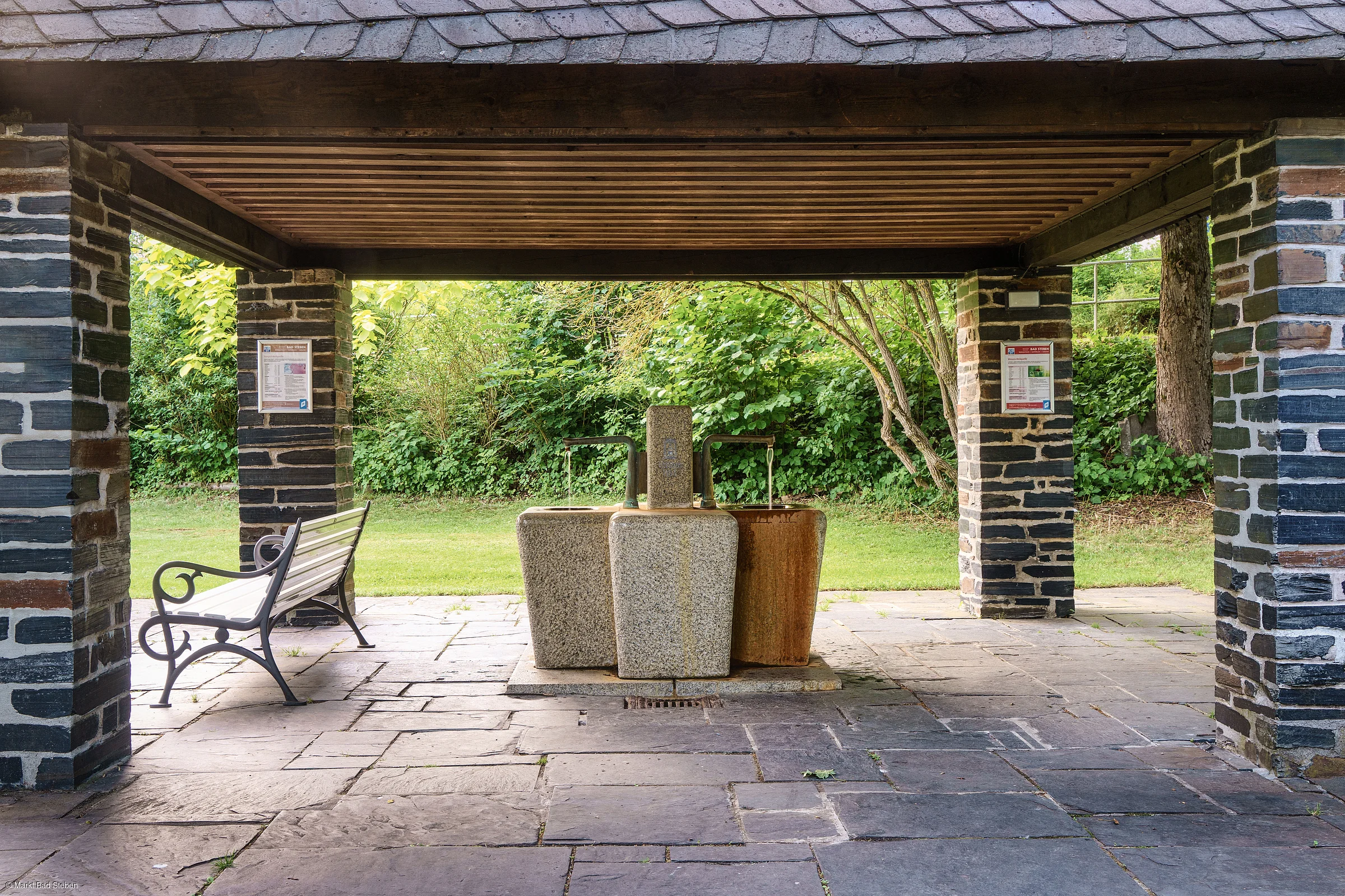 Überdachter Bereich mit drei Wasserbrunnen aus Stein und einer weißen Parkbank, umgeben von Grünflächen.