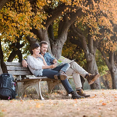 Paar sitzt auf einer Parkbank unter herbstlichen Bäumen und schaut gemeinsam auf eine Karte.