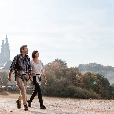 Paar mittleren Alters hält Händchen beim Spaziergang auf einem Weg mit Bäumen und Gebäuden im Hintergrund.