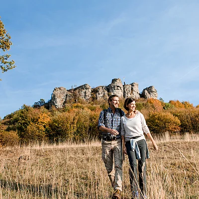 Paar beim Wandern auf einer Wiese mit herbstlichen Bäumen und Felsen im Hintergrund unter blauem Himmel