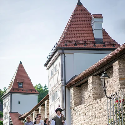 Gruppe mit historisch gekleidetem Stadtführer vor Fachwerkhäusern und Stadtmauer mit Türmen und Laterne.
