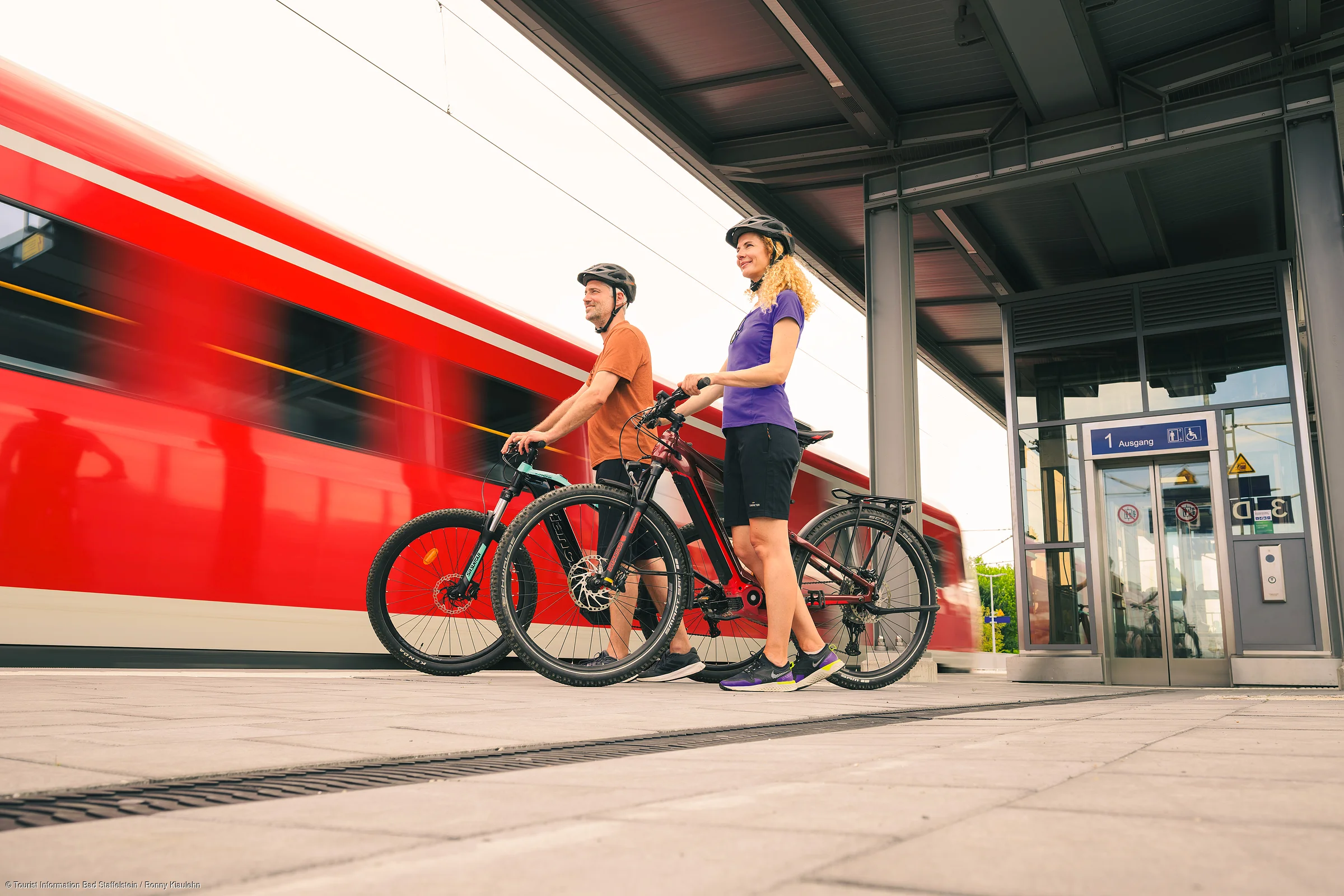 Zwei Radfahrer mit Helmen stehen mit ihren Fahrrädern an einem Bahnsteig, im Hintergrund fährt ein roter Zug vorbei.