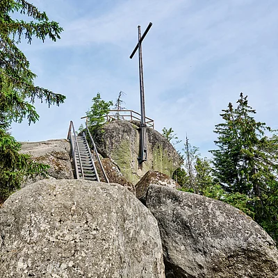 Holzkreuz auf Felsen mit Metalltreppe und umgeben von Nadelbäumen unter blauem Himmel