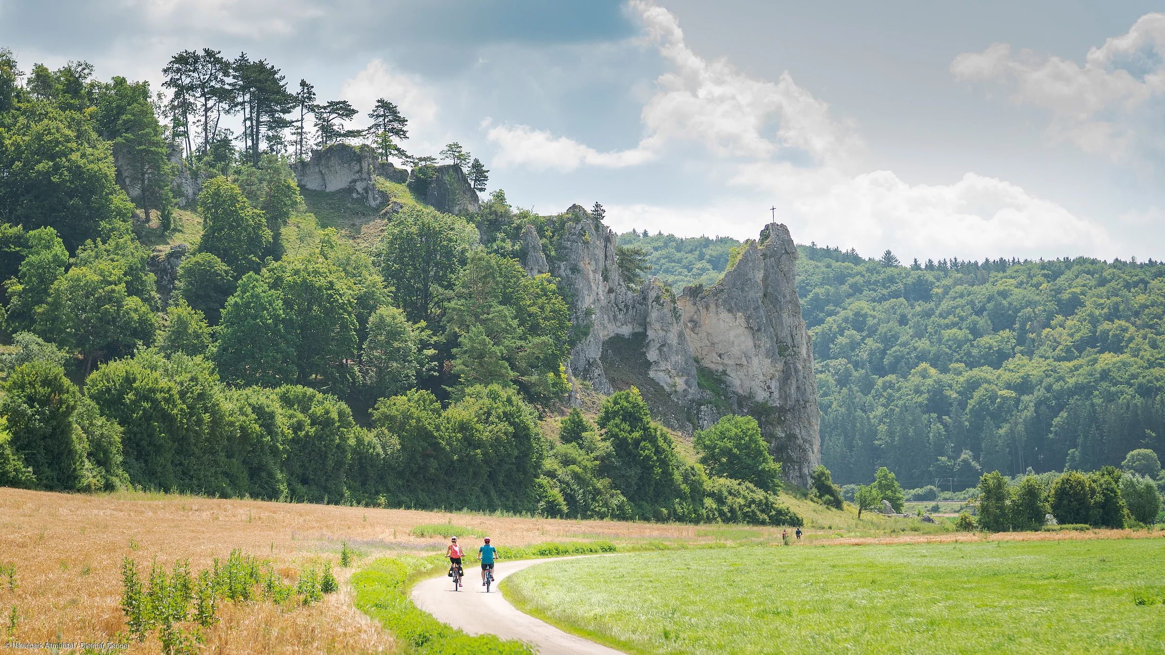 Zwei Radfahrer auf einem Weg durch eine grüne Landschaft mit Felsen und Bäumen im Hintergrund.