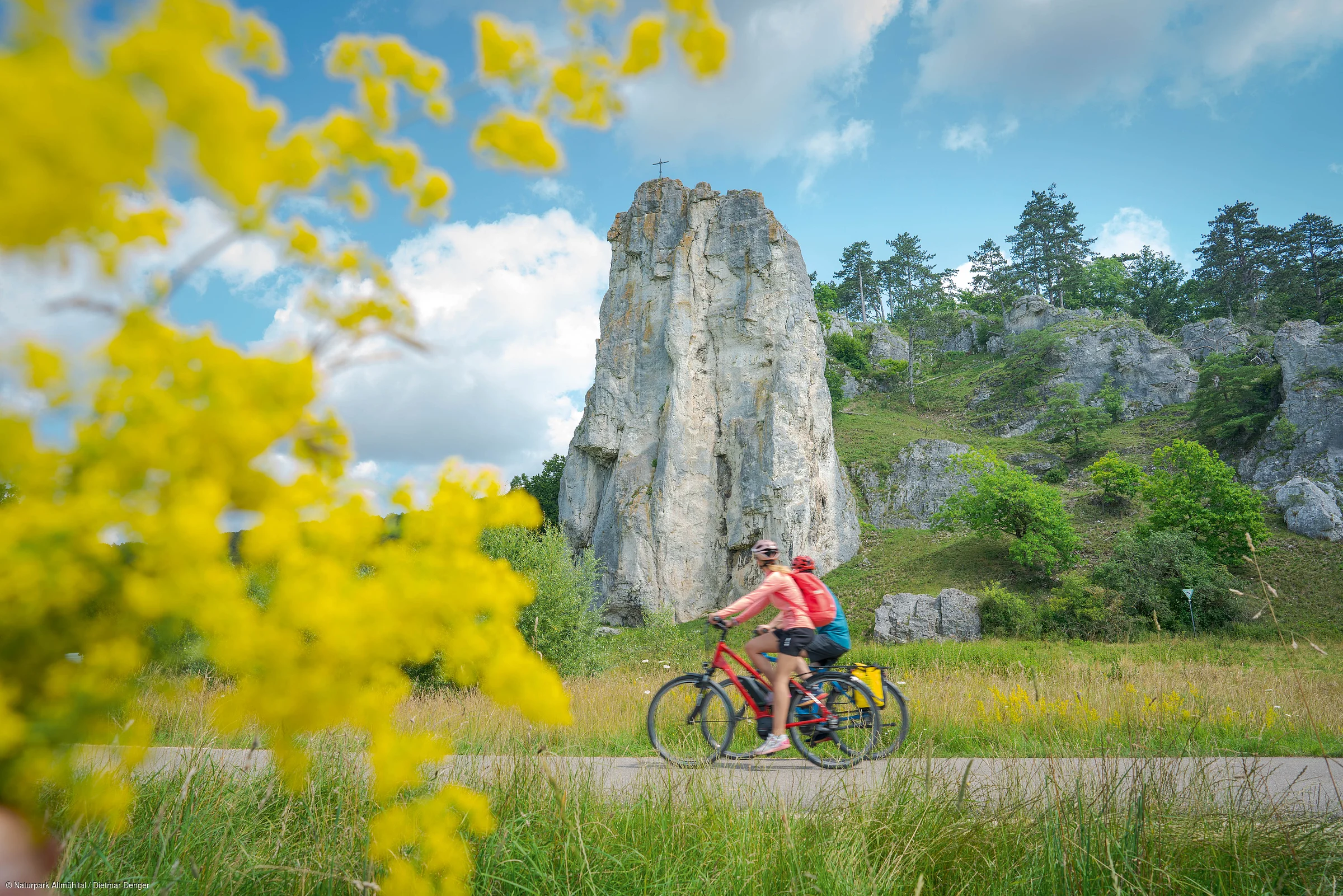 Zwei Radfahrer fahren auf einem Weg vor einem großen Felsen mit Kreuz, umgeben von grüner Landschaft und gelben Blumen.