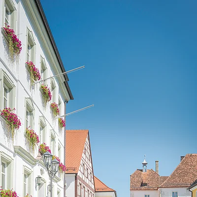 Historische Gebäude mit Blumenkästen und vier Radfahrern auf gepflastertem Platz bei klarem Himmel.