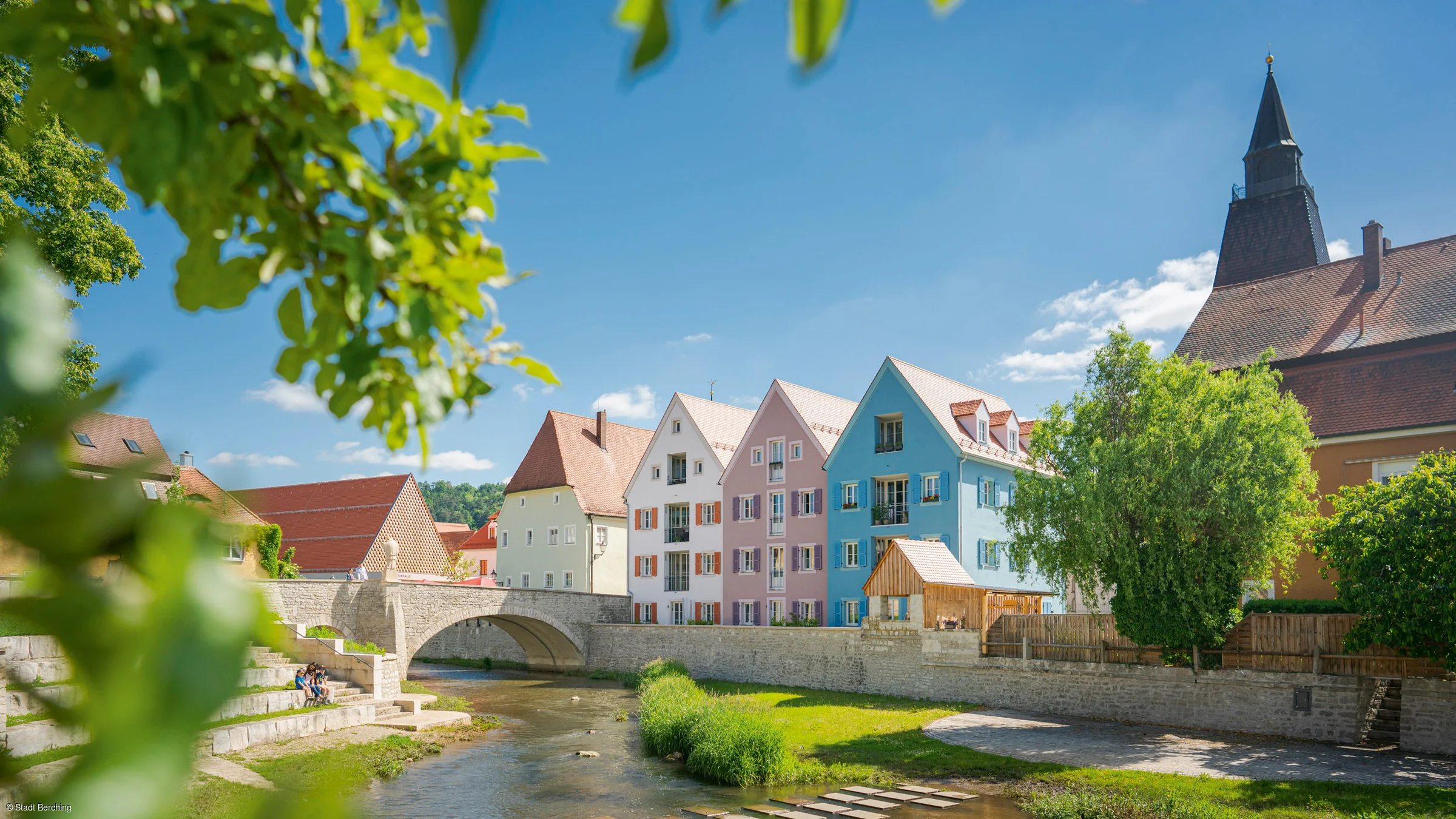 Bunte Häuserreihe an einem Fluss, mit einer Steinbrücke im Vordergrund und einem Kirchturm im Hintergrund.