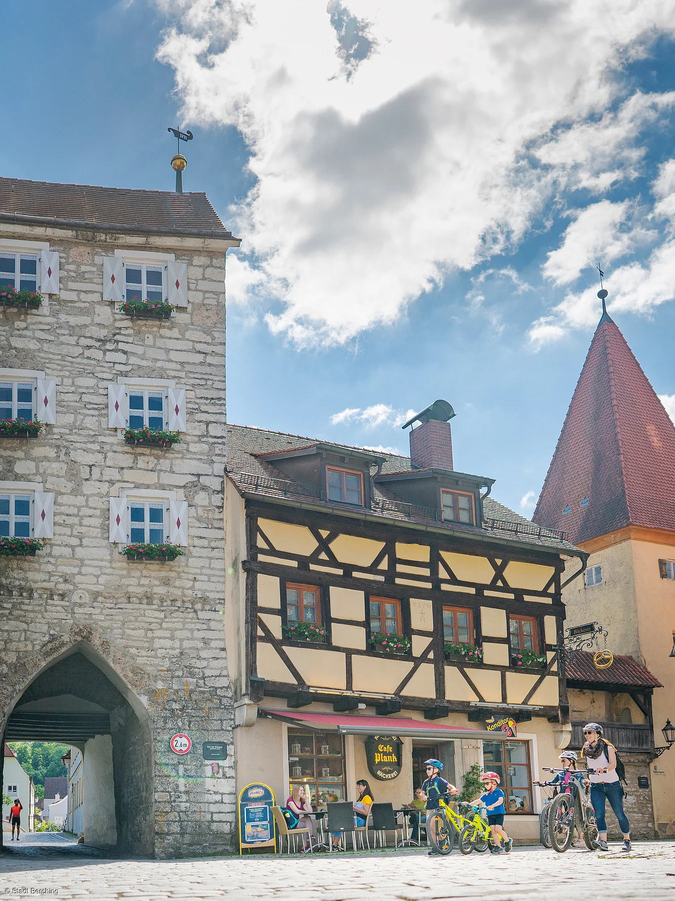 Historisches Gebäude mit Fachwerk und Torbogen, Menschen mit Fahrrädern und Café im Vordergrund.