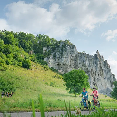 Zwei Radfahrer fahren auf einem Weg vor einer Felsformation und grüner Landschaft.